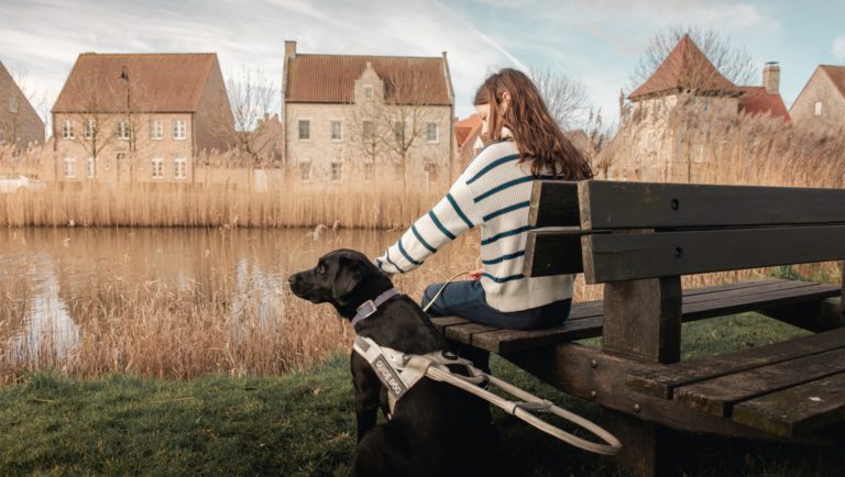 Afbeelding van een jong meisje dat op een houten bank zit naast haar blindengeleidehond. Ze bevinden zich voor een meer met hoge grassen en huizen op de achtergrond. Het meisje aait haar hond die een harnas draagt. We zien de rug van het meisje en de hond die genieten van het moment.